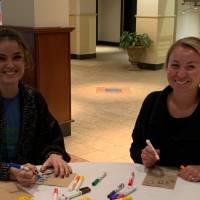 Two women sitting at a table decorating lunch bags for Kids' Food Basket